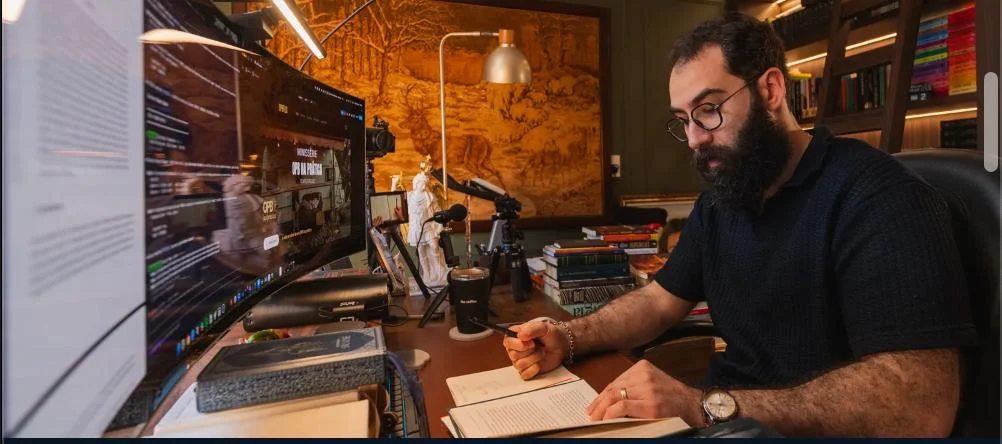 Elton Luiz escrevendo em seu escritório, cercado por livros e equipamentos de produção